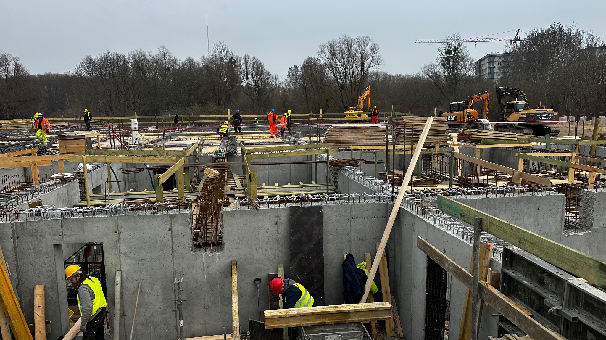 Budowa fundamentów domu w trakcie realizacji: widoczne zbrojenia, szalunki, betonowe ściany, pracownicy w kaskach i kamizelkach odblaskowych, oraz maszyny budowlane w tle.