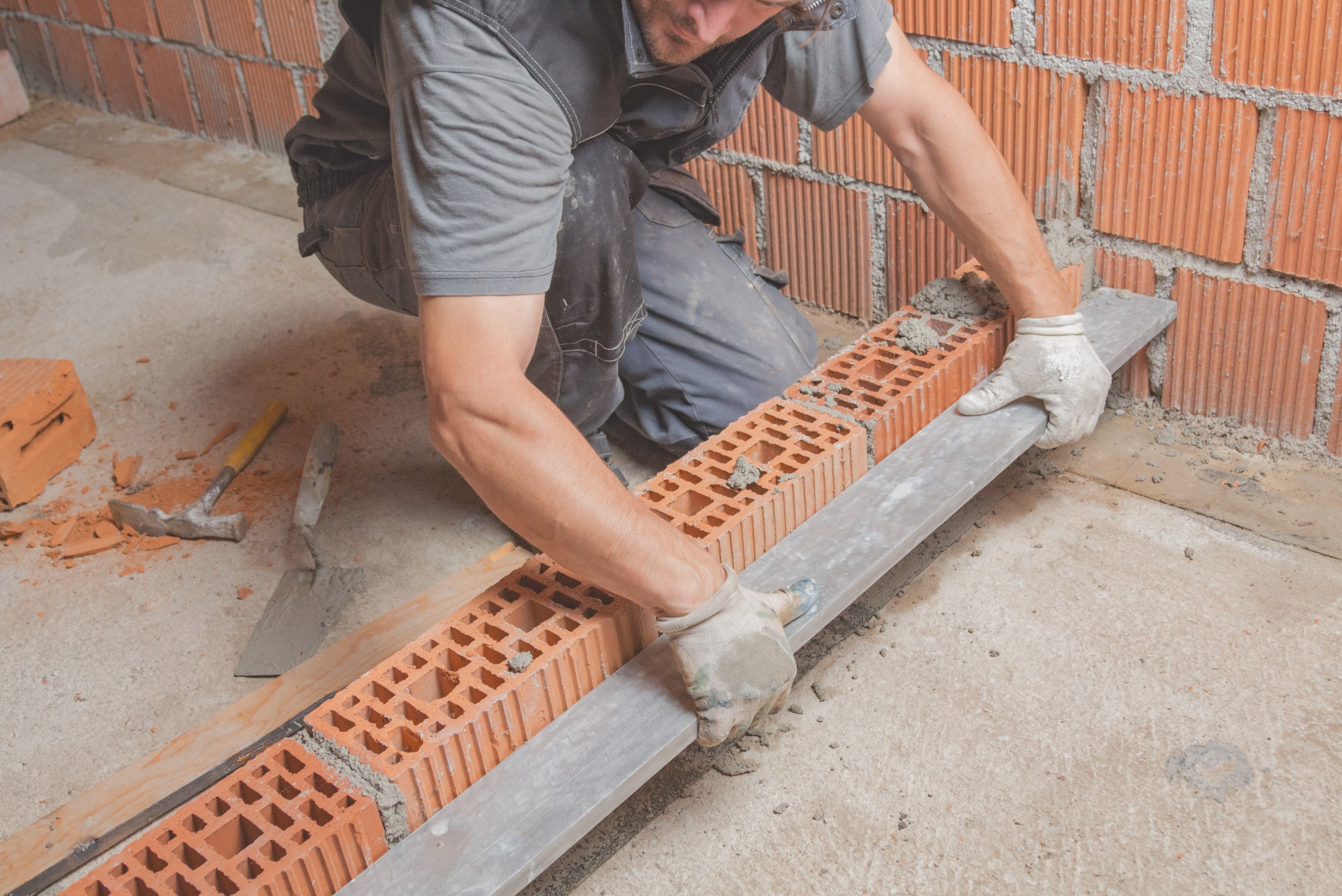 Murarz w trakcie pracy: układa bloczki z betonu komórkowego na zaprawie, wyrównując je łatą. W tle widoczna ściana z czerwonej cegły dziurawki.