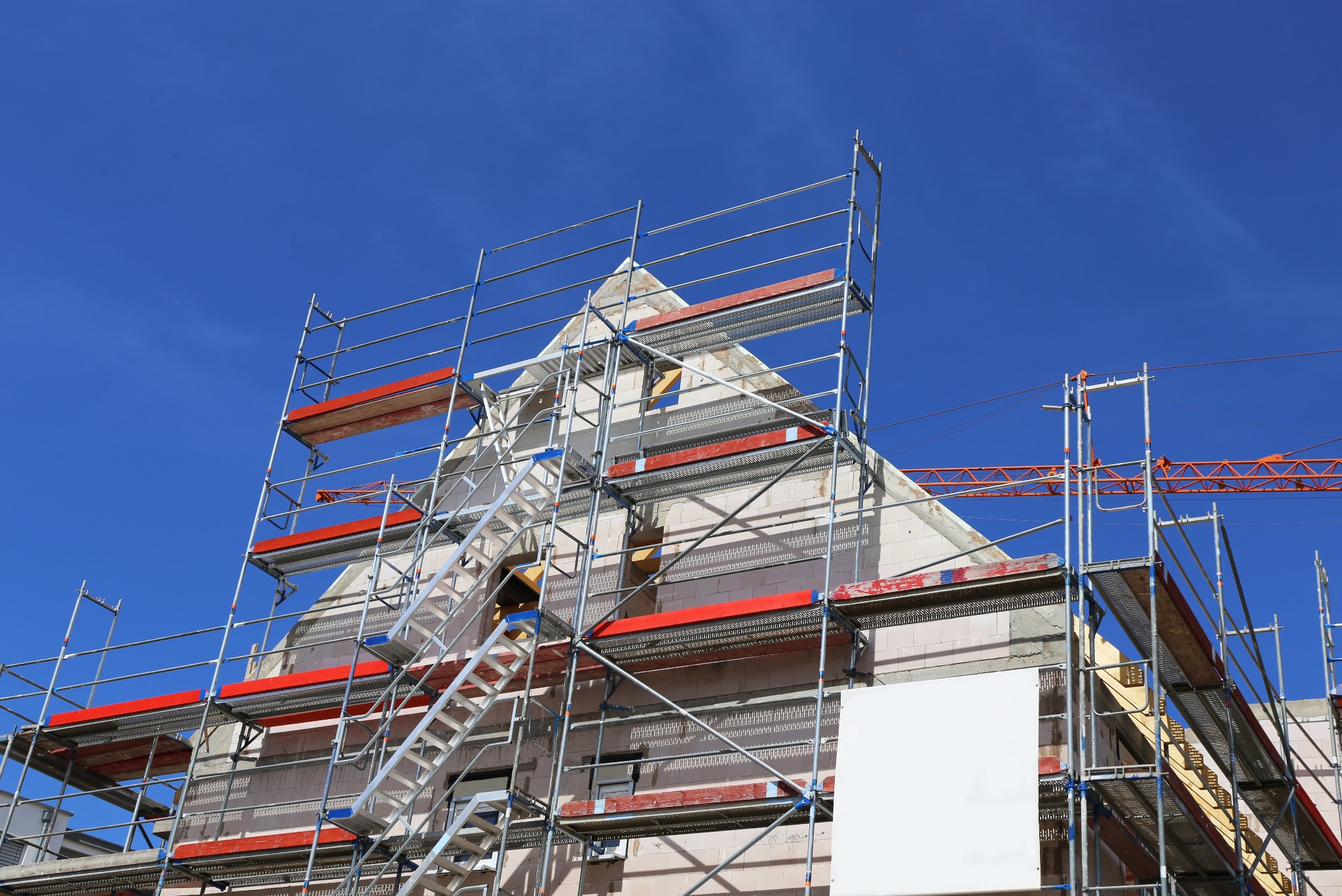 Budowa domu z białej cegły, otoczona rusztowaniami na tle błękitnego nieba. Widoczny fragment żurawia budowlanego w tle. Ujęcie z dołu.