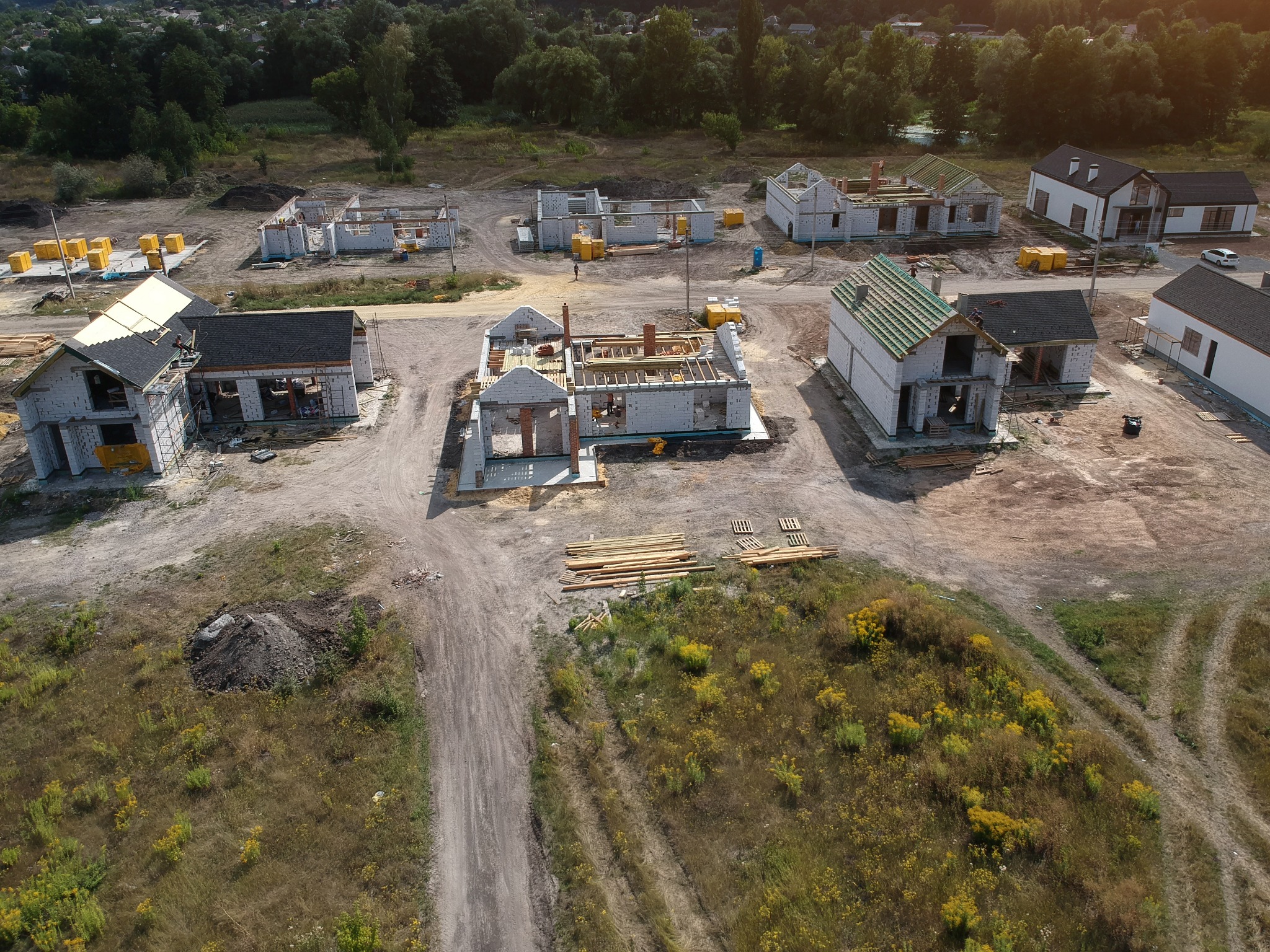 Osiedle w budowie z lotu ptaka: domy w różnym stadium zaawansowania, od fundamentów po gotowe elewacje. Widoczne materiały budowlane i otaczający teren.