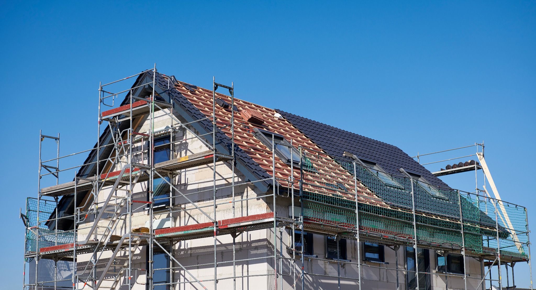 Dom w trakcie krycia dachu, widoczne rusztowania i część dachu pokryta czerwoną dachówką, a część czarną. Na dachu okna dachowe. Błękitne niebo w tle.