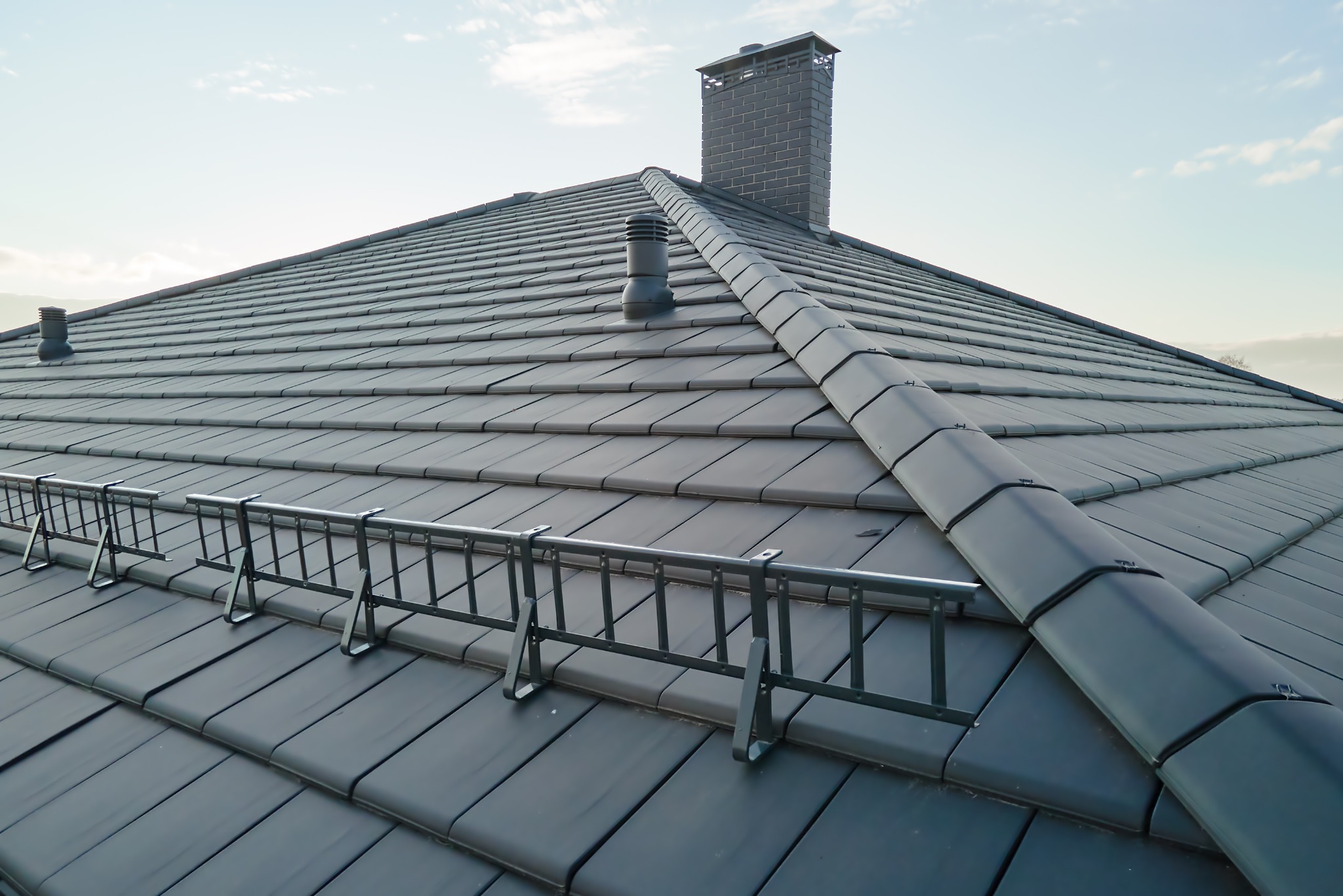 Grafitowy dach z ceramicznej dachówki karpiówki, wyposażony w system komunikacji dachowej i komin z cegły klinkierowej na tle jasnego nieba.
