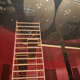 FHU Akarw Adam karwowski - Pracownik na rusztowaniu czyści sufit z okrągłymi elementami dekoracyjnymi w pomieszczeniu z czerwoną ścianą. Widok z dołu, perspektywa zbieżna.