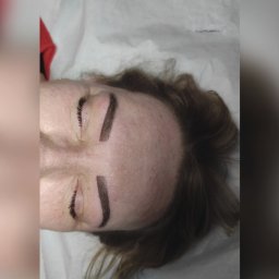 Permanent Makeup Kinga Budziak - Widok z góry na twarz kobiety z nowo wykonanym makijażem permanentnym brwi, leżącej na białym podkładzie.