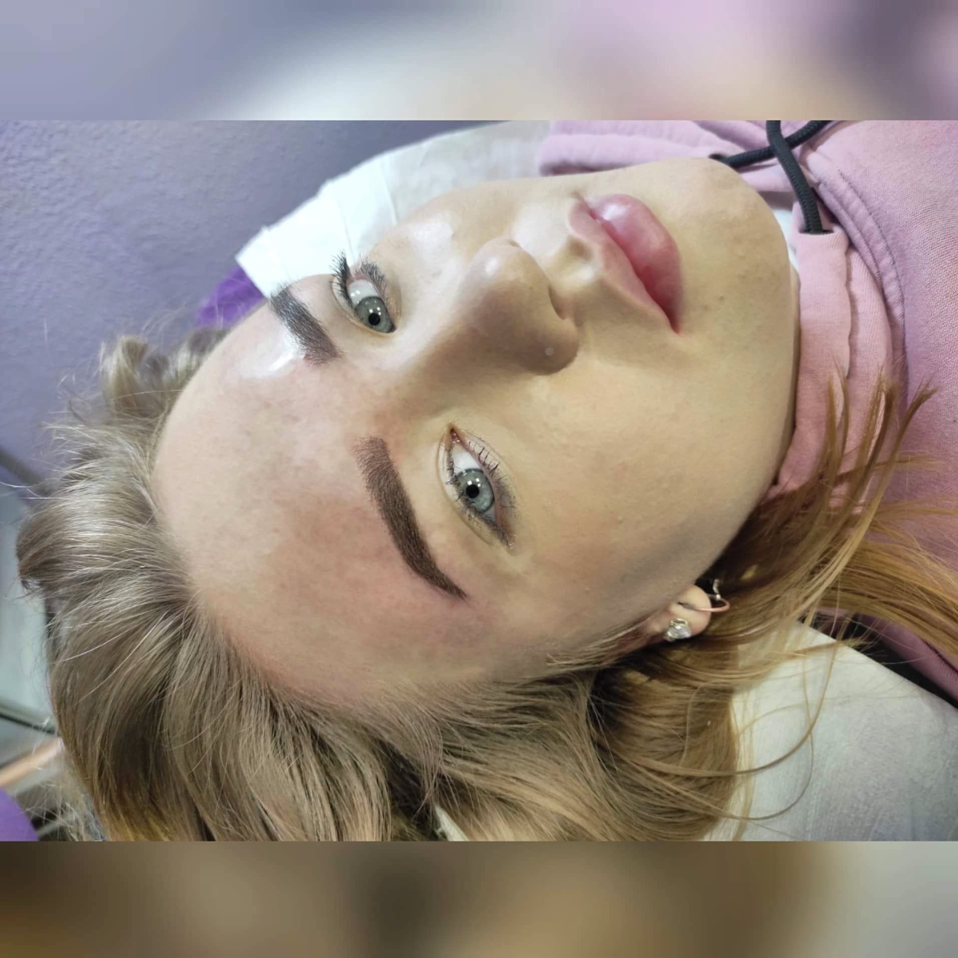 Blondynka z delikatnym makijażem permanentnym brwi, leżąca na białym podłożu, widoczne kolczyki w uszach.