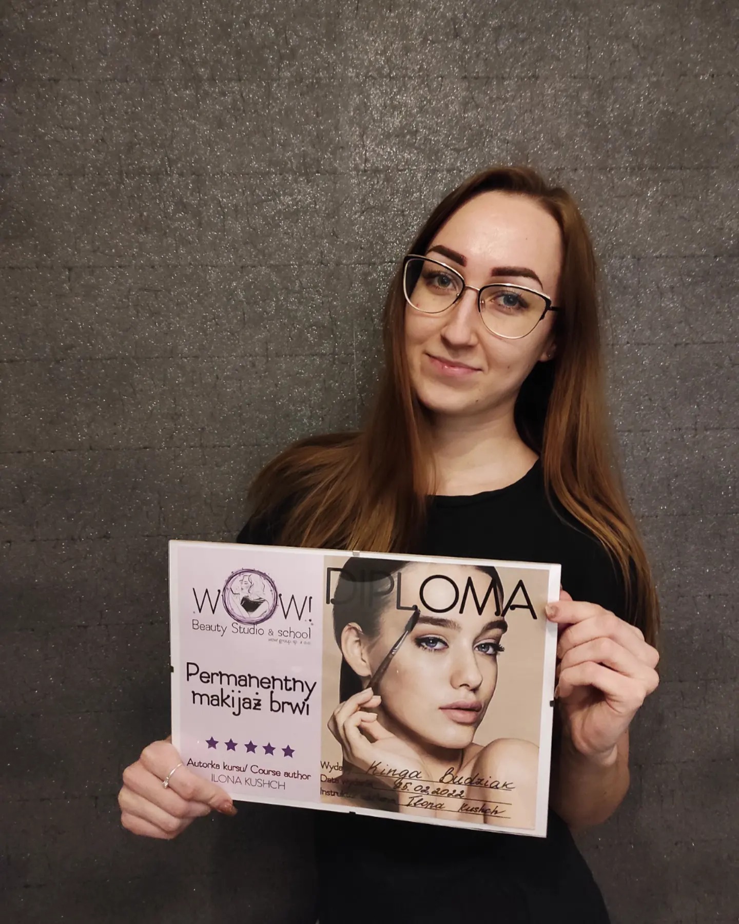 Kobieta w okularach prezentuje dyplom ukończenia kursu permanentnego makijażu brwi, z logo 'WOW! Beauty Studio & school' i zdjęciem modelki z wykonanym makijażem brwi.