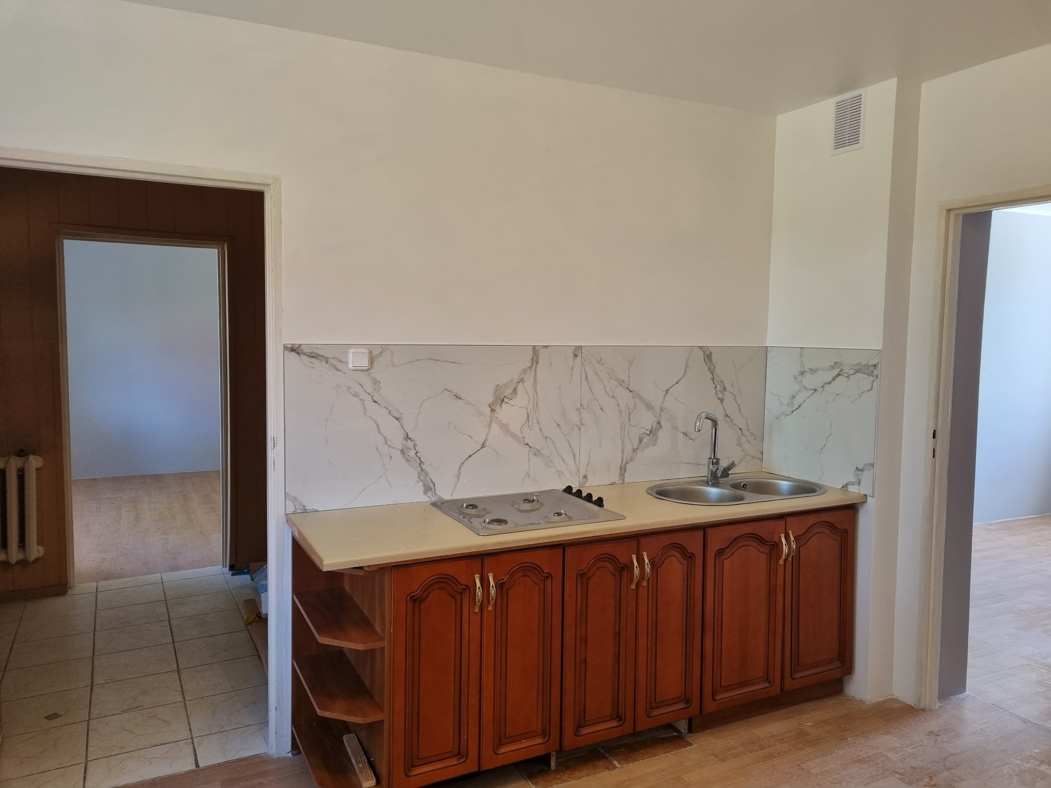 Kuchnia po remoncie: zabudowa z brązowymi szafkami, marmurowe panele nad blatem, widoczna kuchenka gazowa i zlewozmywak dwukomorowy, podłoga wyłożona panelami.