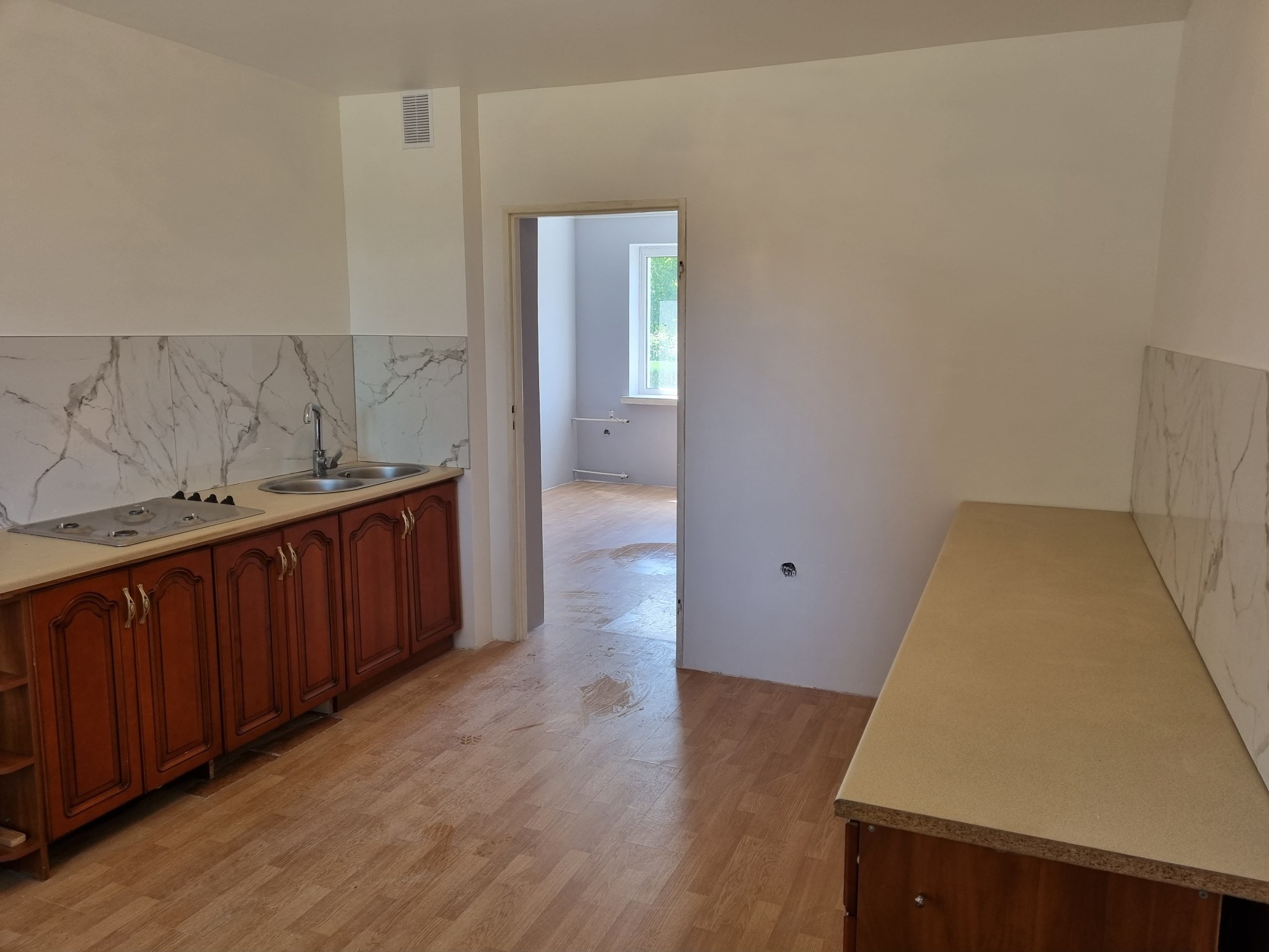 Kuchnia po remoncie z drewnianymi szafkami, marmurowymi płytkami nad blatem i świeżo pomalowanymi ścianami na biało; widoczne ślady po remoncie na podłodze z paneli.