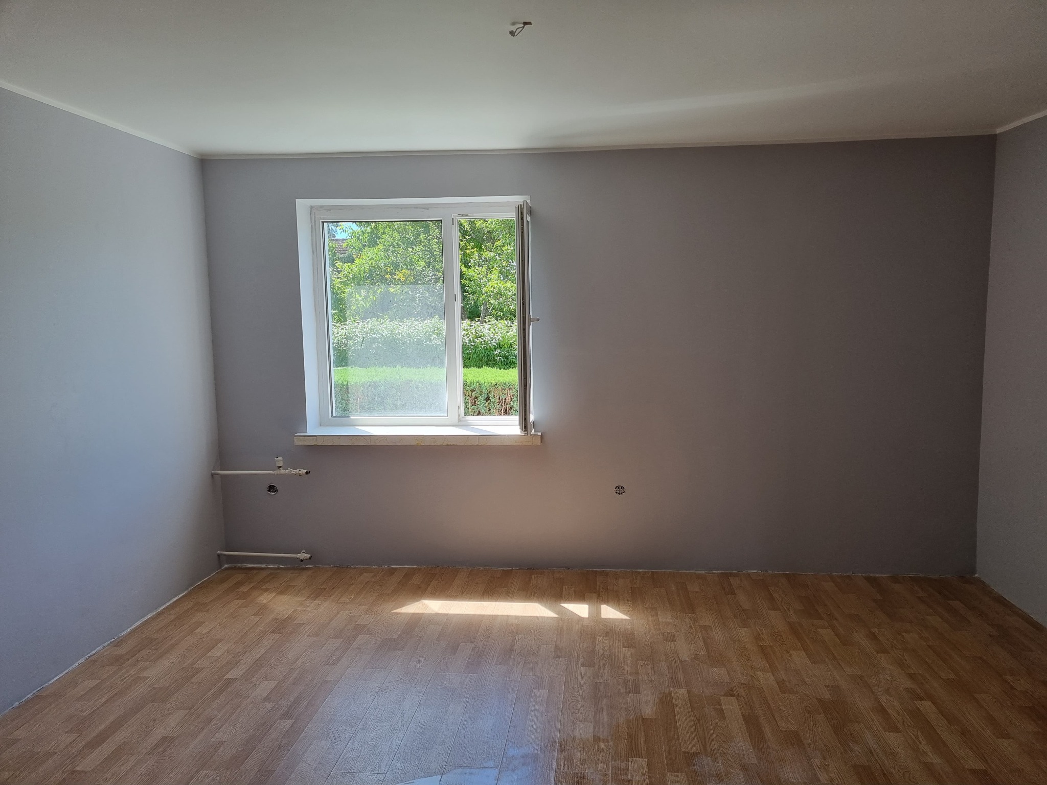 Pusty pokój po remoncie z nową podłogą z paneli imitujących drewno, szare ściany i białym oknem z widokiem na zieleń, widoczne rury grzewcze przy ścianie.