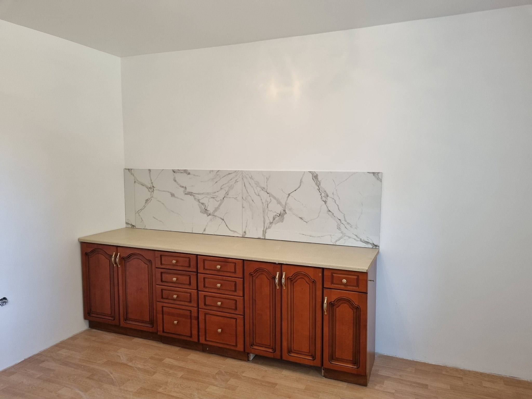 Kuchnia po remoncie: klasyczne, ciemne szafki z beżowym blatem i panelem ściennym imitującym biały marmur, na podłodze panele imitujące drewno.