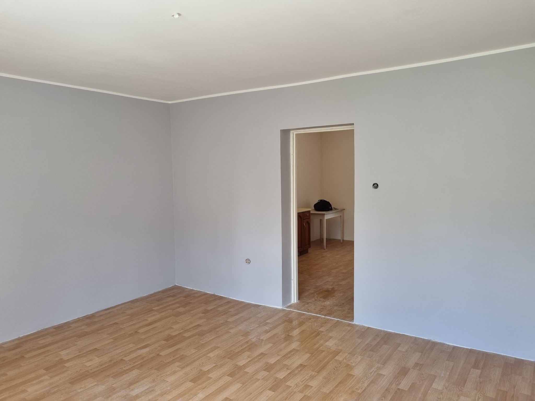 Pomalowane na szaro wnętrze pokoju z panelami podłogowymi, widok na sąsiednie pomieszczenie z meblami.