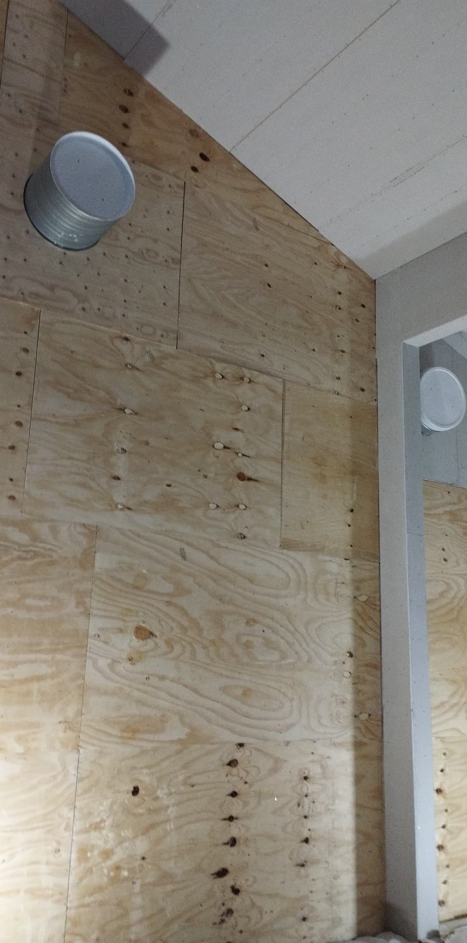 Ściana z surowej sklejki z widocznymi śladami po wkrętach i okrągłym otworem wentylacyjnym, fragment sufitu i narożnika ściany z ościeżnicą w tle.