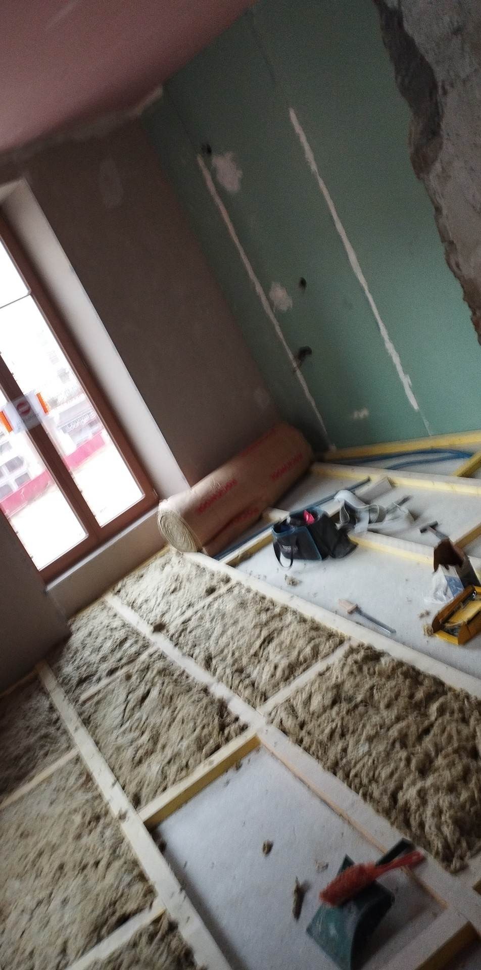 Izolacja podłogi wełną mineralną w trakcie prac, widoczne drewniane legary, rolka materiału izolacyjnego, narzędzia i torba robocza przy ścianie, surowy stan ścian.