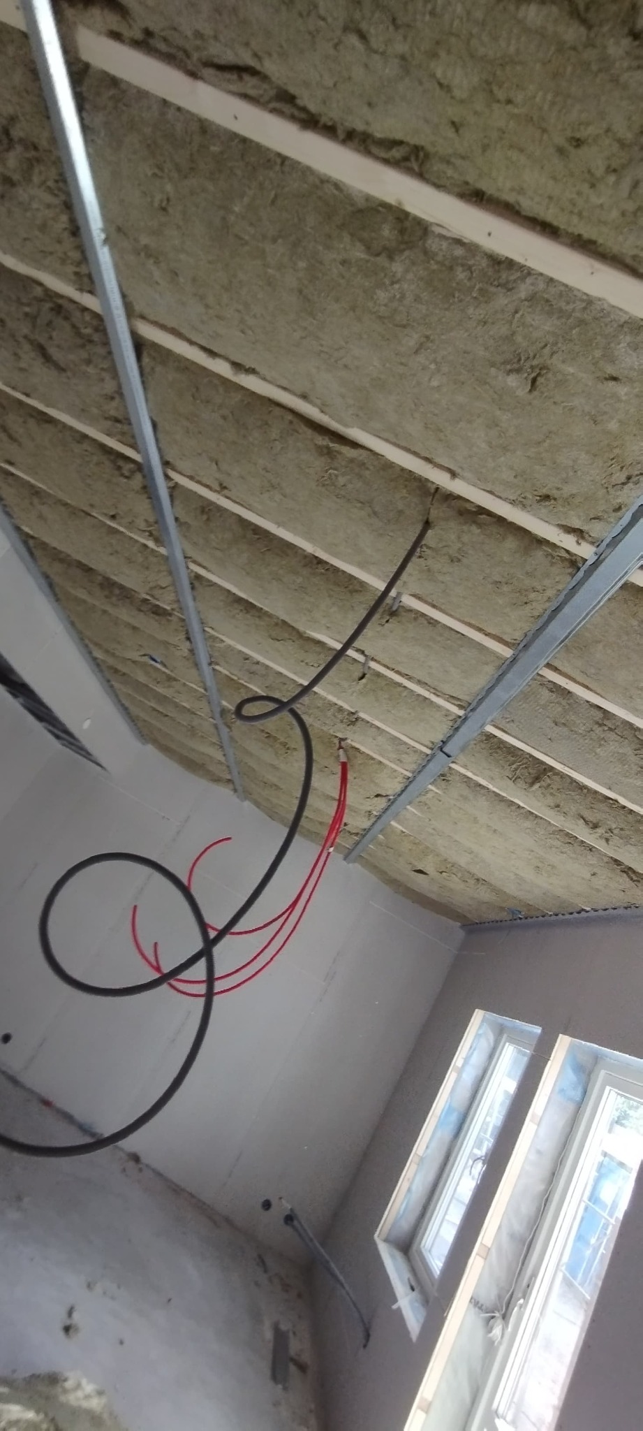 Izolacja z wełny mineralnej zamontowana pod konstrukcją sufitu podwieszanego, widoczne metalowe profile i przewody elektryczne oraz instalacja wodna w nowo wykończonym pomieszczeniu z oknami.