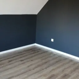 Pomieszczenie na poddaszu z ukośną ścianą pomalowaną na granatowo, widoczna podłoga z paneli i wiadro po farbie w rogu.