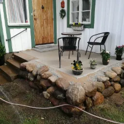 Umyta wodą kamienna podmurówka tarasu z meblami ogrodowymi, schodami i kwiatami w donicach przed domem z drewnianą elewacją.