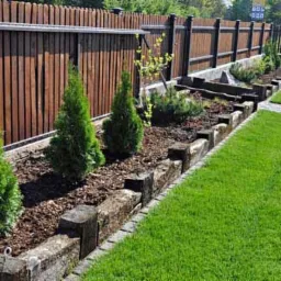 Ogród z równo przystrzyżonym trawnikiem, obrzeżami z drewnianych bali i rządkiem posadzonych, młodych tuj przy drewnianym płocie.