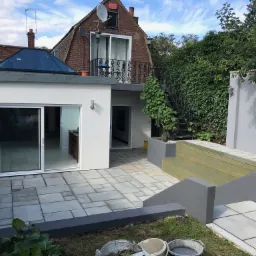 Wykończony taras z szarych płyt, przylegający do białego budynku z przeszklonym dachem, z metalowymi schodami prowadzącymi na balkon na piętrze, widoczne wiaderka po farbie na trawniku.