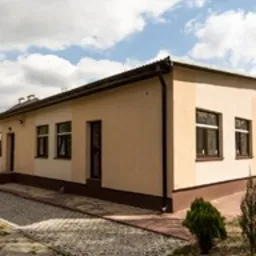 Budynek parterowy z beżową elewacją, brązowymi oknami i drzwiami, widoczny chodnik z kostki brukowej i fragment trawnika. Dach dwuspadowy, jasne niebo z lekkimi chmurami.