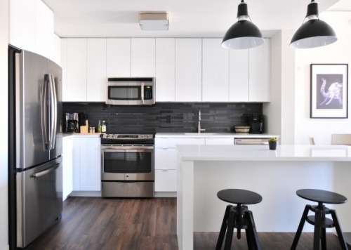 Nowoczesna kuchnia z białymi szafkami, ciemnoszarym backsplashiem z płytek, stalowym sprzętem AGD i ciemną podłogą z desek. Dwie czarne, regulowane wysokością stołki barowe przed białą wyspą...