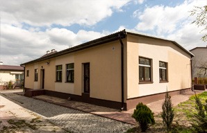 Budynek parterowy z beżową elewacją, brązowymi oknami i drzwiami, widoczny chodnik z kostki brukowej i fragment trawnika. Dach dwuspadowy, jasne niebo z lekkimi chmurami.