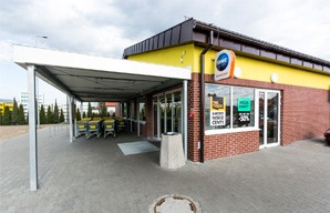 Zewnętrzny widok sklepu z żółtą elewacją, ceglanym cokołem i zadaszonym wózkiem na zakupy przy wejściu.