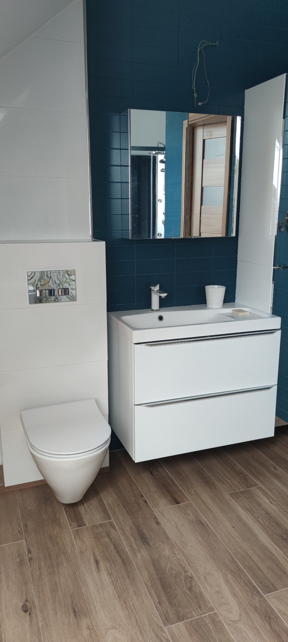 Nowoczesna łazienka z białymi meblami, toaletą i umywalką na tle ciemnoniebieskich płytek, z drewnopodobną podłogą i widocznymi przewodami elektrycznymi.