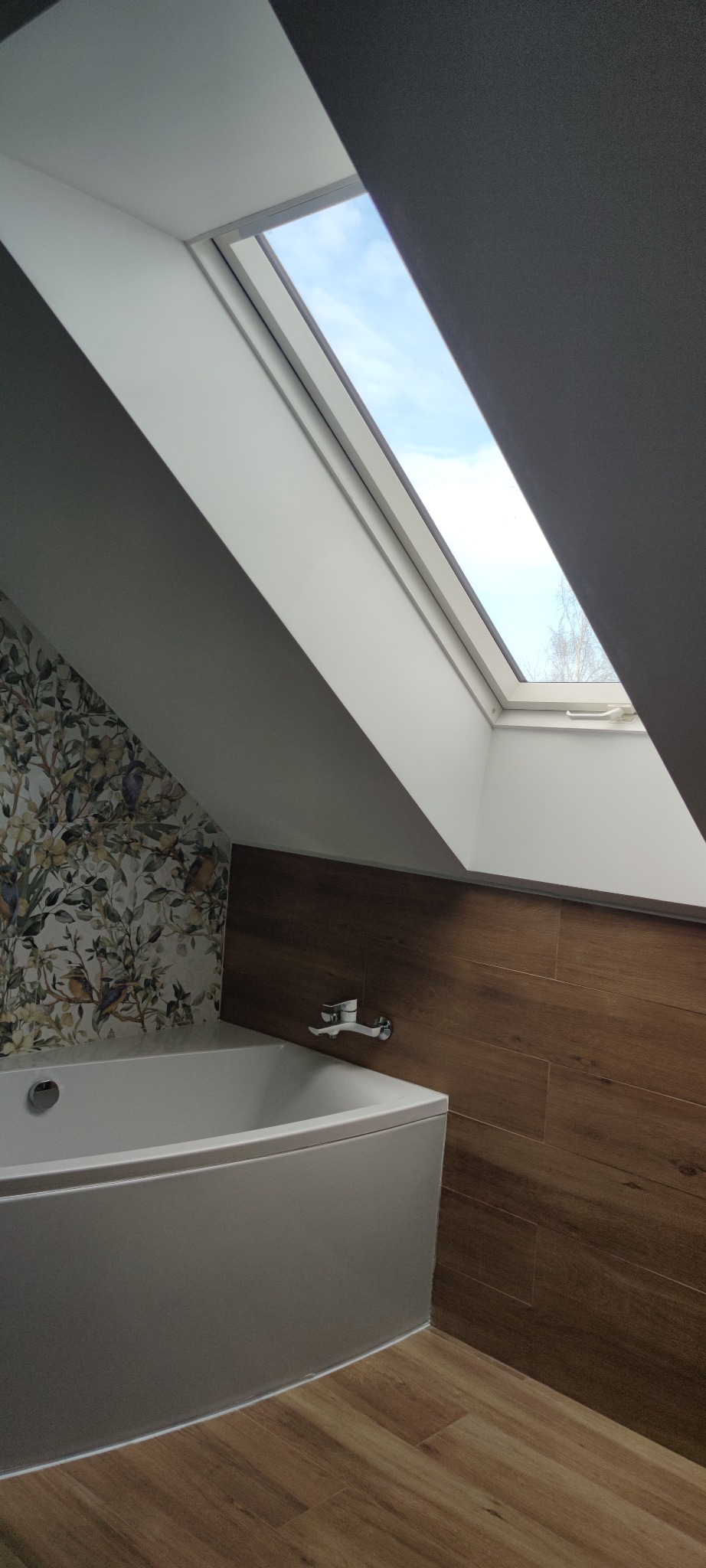 Biała wanna w łazience na poddaszu z oknem dachowym, wykończona płytkami imitującymi drewno i tapetą w motywy roślinne, bateria umieszczona na ścianie.