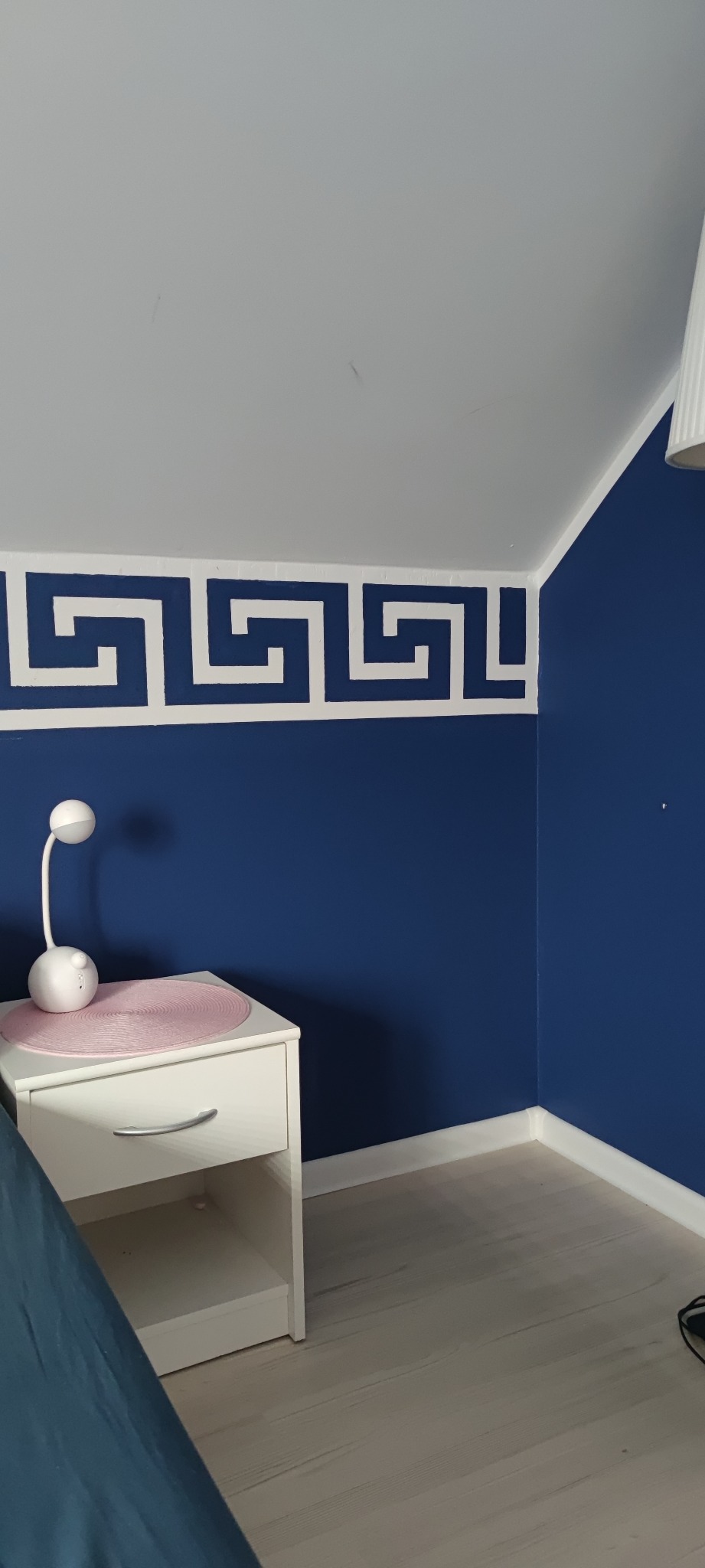 Granatowa ściana z białym greckim wzorem malowanym pod sufitem, biała szafka nocna z lampką, drewniana podłoga w pokoju na poddaszu.