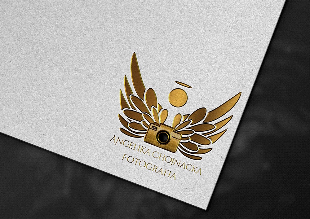 Złote logo firmy fotograficznej: aparat z rozłożonymi skrzydłami anioła i aureolą nad nim, umieszczone na teksturowanym, białym papierze z cieniem.