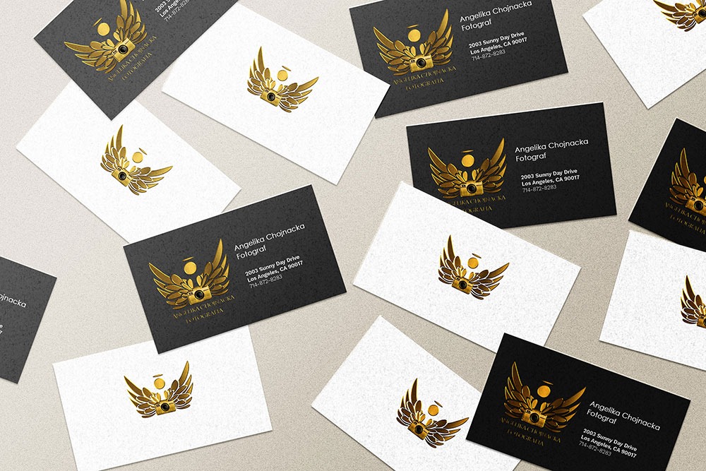 Rozłożone wizytówki fotografa na białym i czarnym tle z złotym logo przedstawiającym aparat z anielskimi skrzydłami.