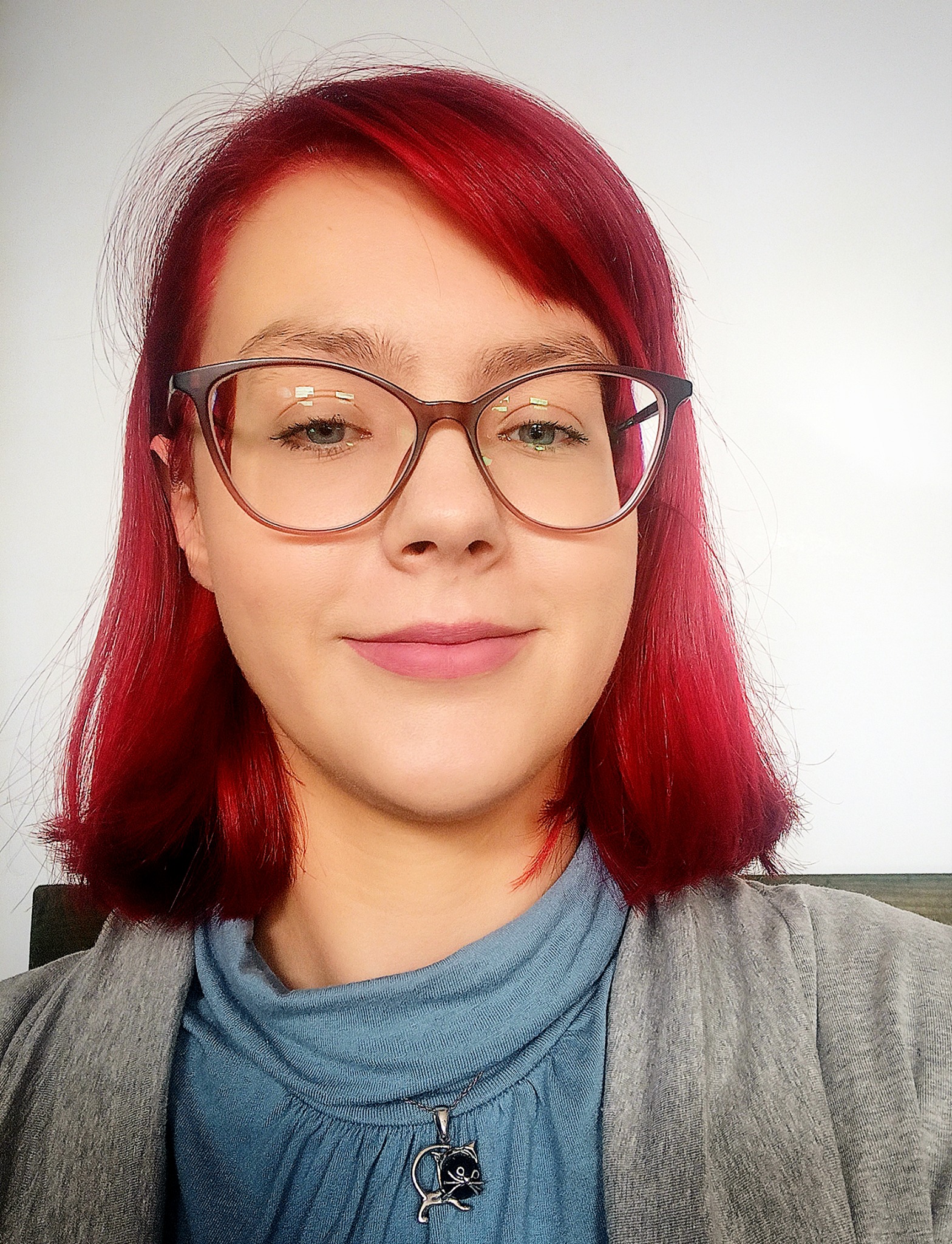 Portret kobiety z czerwonymi włosami w okularach, uśmiechniętej, z wisiorkiem w kształcie kota.