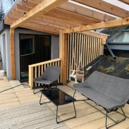 Zadaszony taras z drewnianą podłogą, szarą sofą i fotelem oraz czarnym stolikiem kawowym, obok huśtawka dla dziecka zawieszona na drewnianej konstrukcji, w tle okno dachowe.