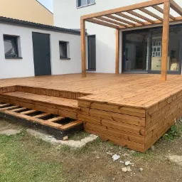 Nowoczesny taras z drewna z pergolą, częściowo wykończony, widoczne elementy konstrukcyjne schodów i podbudowy, na tle białego budynku z szarymi oknami.