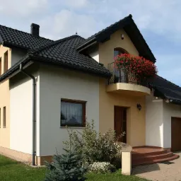 Dwukondygnacyjny dom z ocieploną elewacją w odcieniach beżu i bieli, z czarnym dachem i balkonem ozdobionym czerwonymi kwiatami.