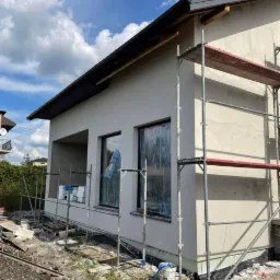Dom jednorodzinny w trakcie nakładania jasnego tynku elewacyjnego, okna zabezpieczone folią, widoczne rusztowanie i przygotowane materiały budowlane.