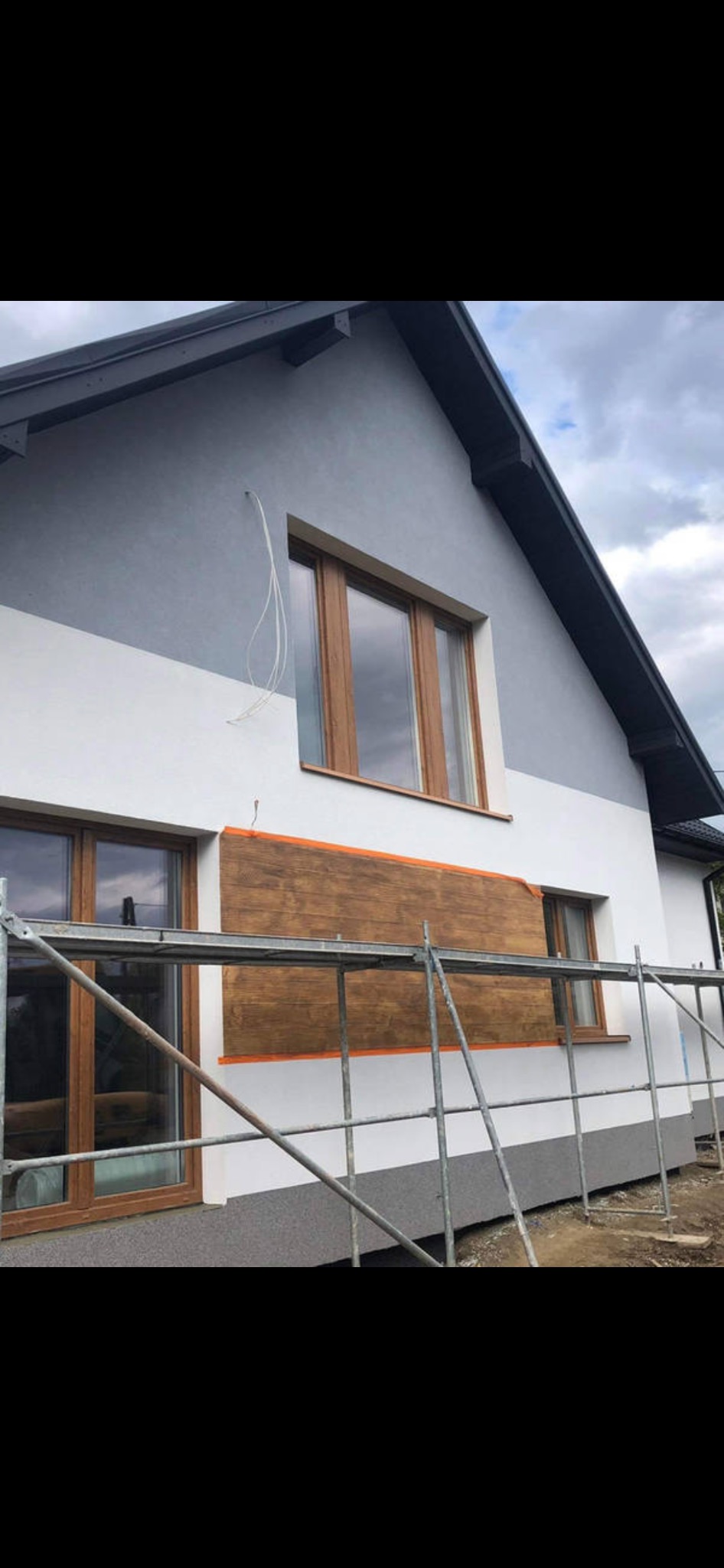 Dwukondygnacyjny dom z fragmentem elewacji zabezpieczonym brązową płytą imitującą drewno i pomarańczową taśmą malarską, widoczne rusztowanie.