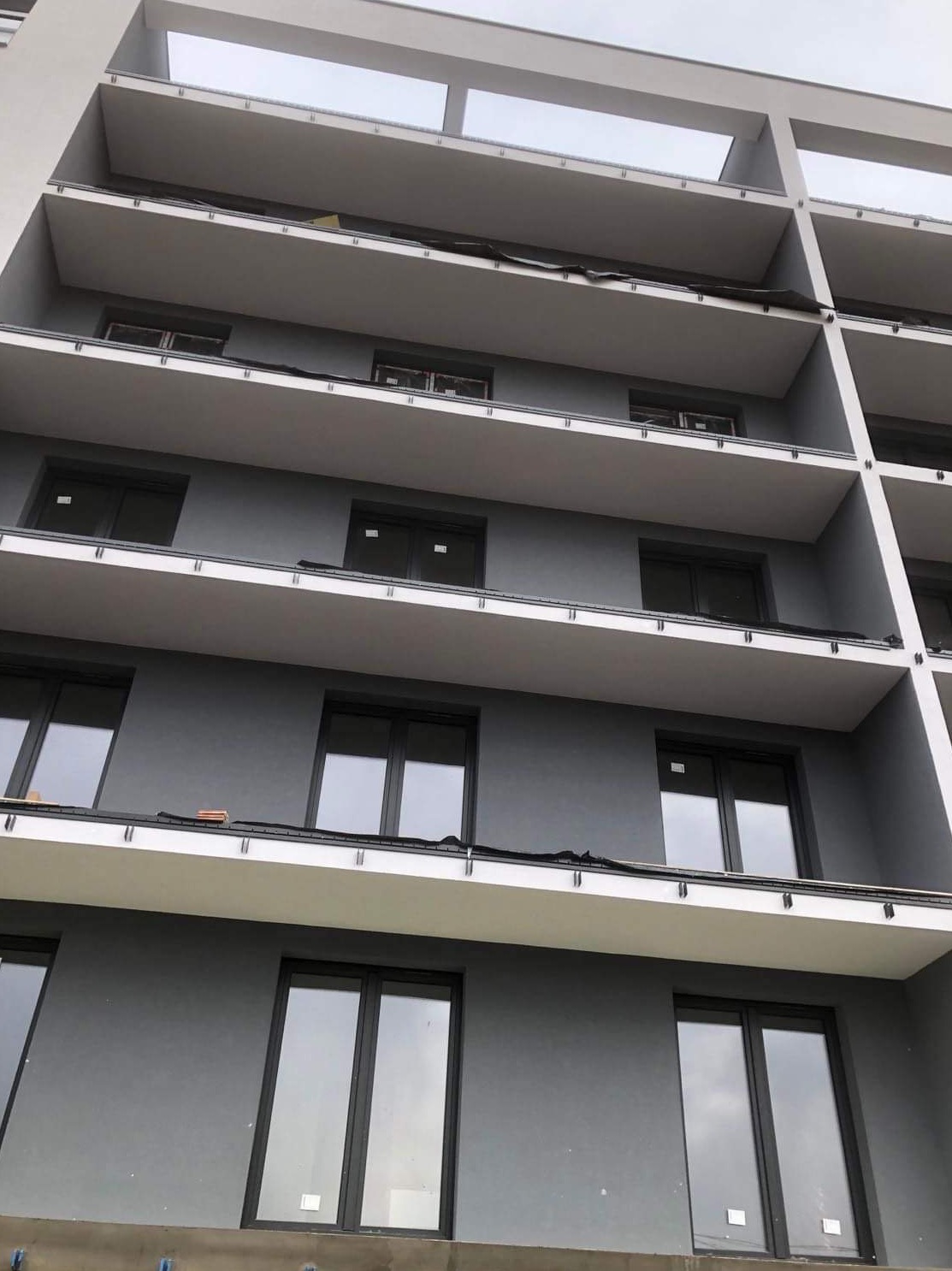 Wielopiętrowy budynek z szarą elewacją i balkonami zabezpieczonymi czarną folią przed dalszymi pracami wykończeniowymi.