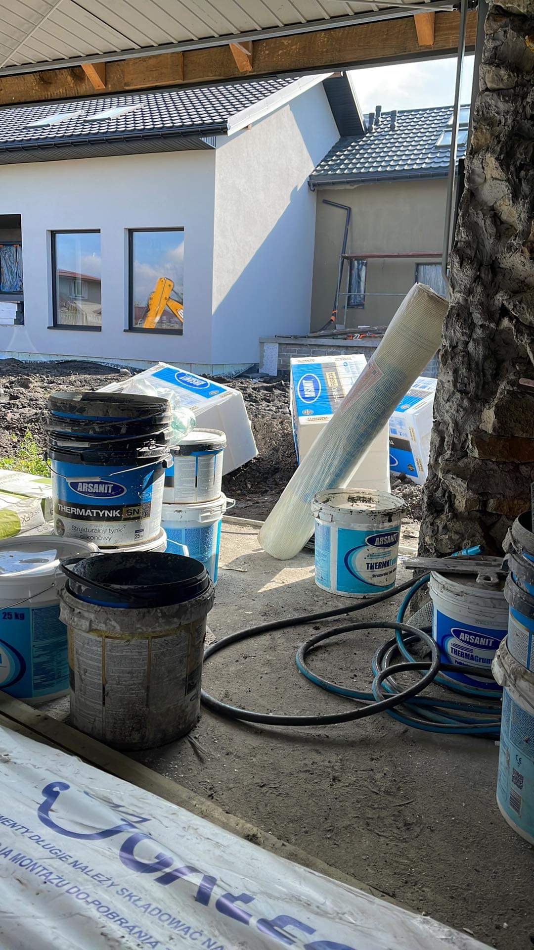 Stos wiader z farbą i tynkiem strukturalnym marki Arsanit na placu budowy, rolka siatki elewacyjnej oparta o ścianę, w tle fragment nowo otynkowanej elewacji domu jednorodzinnego z widocznym...