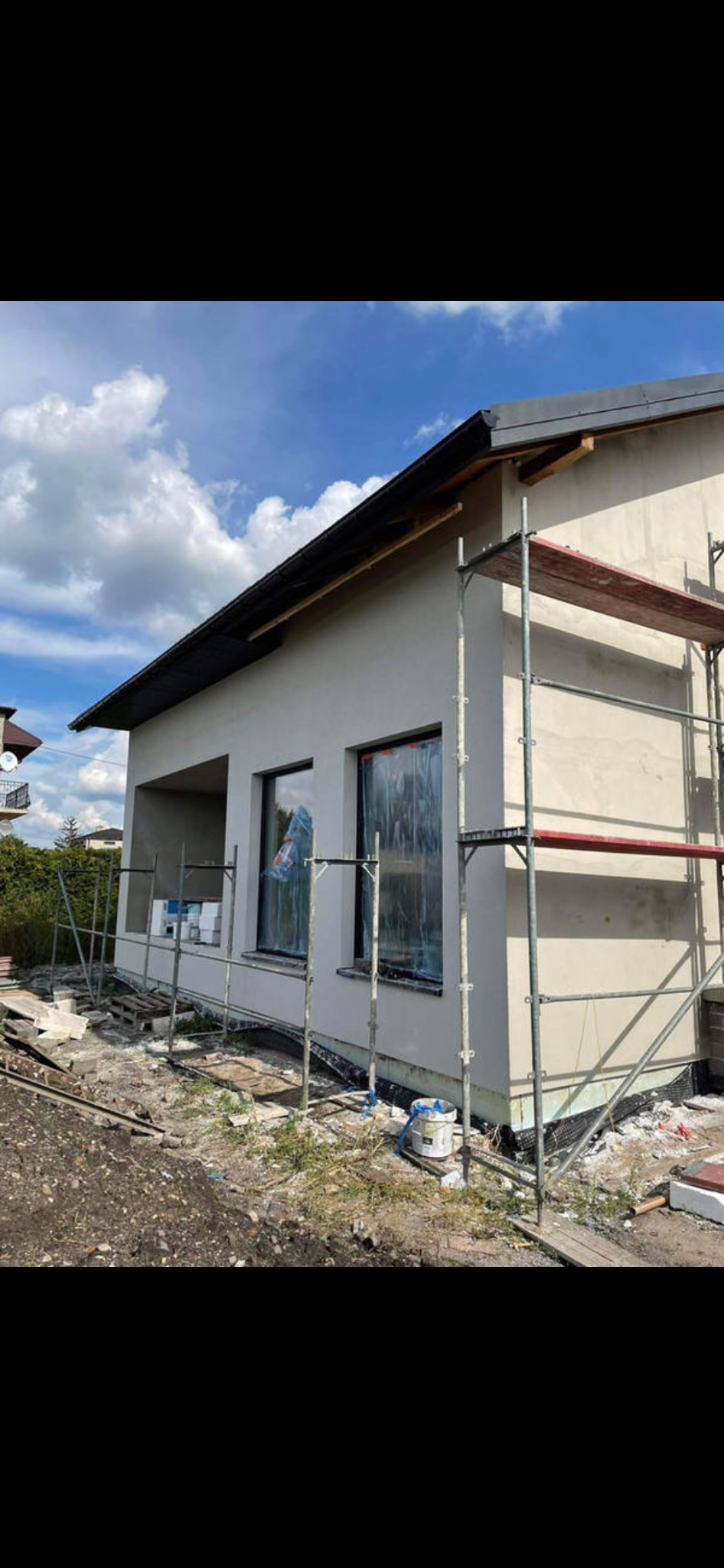 Dom jednorodzinny w trakcie nakładania jasnego tynku elewacyjnego, okna zabezpieczone folią, widoczne rusztowanie i przygotowane materiały budowlane.