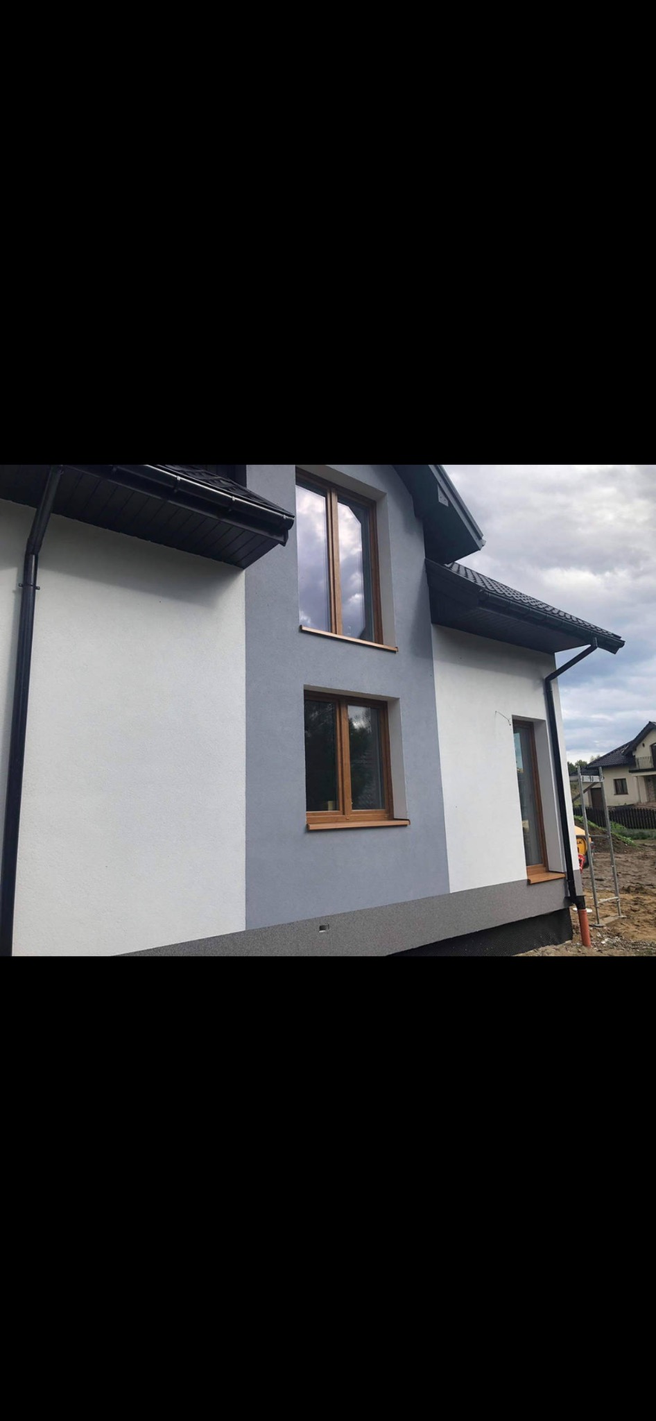 Dwukolorowa elewacja domu jednorodzinnego: biała i szara, z drewnianymi oknami i czarnymi rynnami, widok z boku na tle pochmurnego nieba.