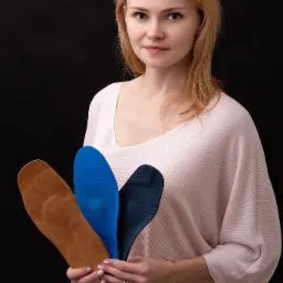 Kobieta prezentuje trzy różne wkładki ortopedyczne: brązową skórzaną, niebieską perforowaną i granatową perforowaną, na czarnym tle.