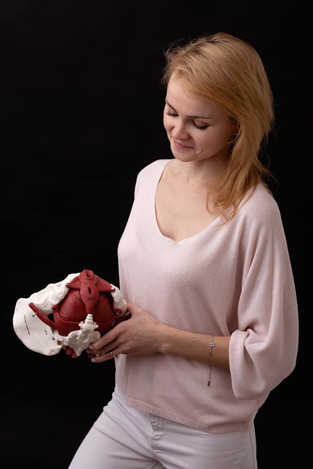 Kobieta w jasnym swetrze trzyma model miednicy z mięśniami dna miednicy na czarnym tle.