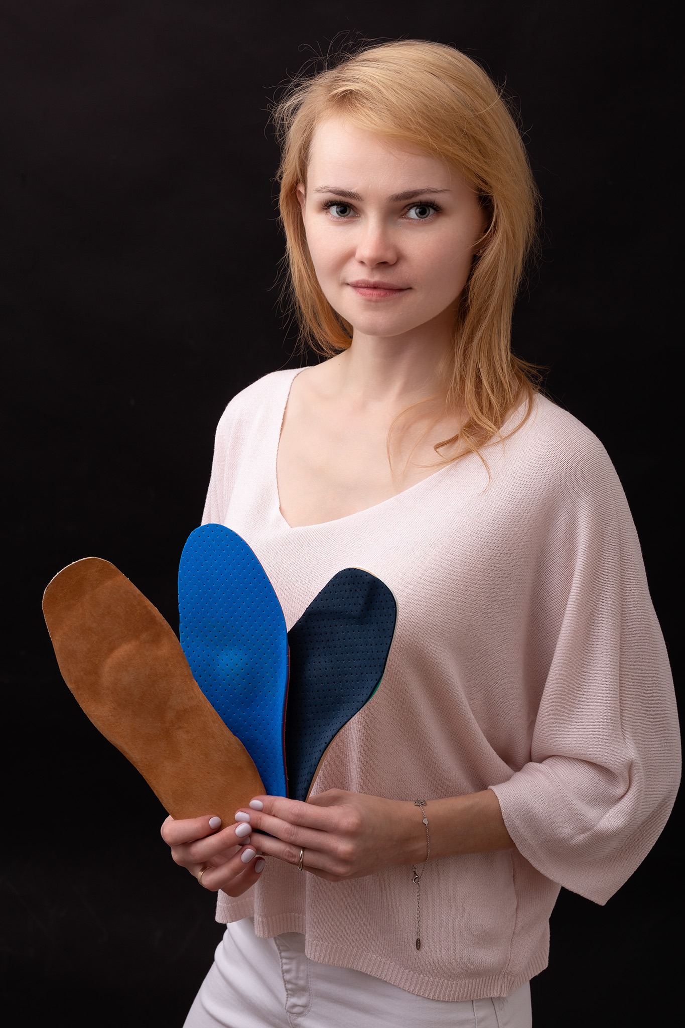 Kobieta prezentuje trzy różne wkładki ortopedyczne: brązową skórzaną, niebieską perforowaną i granatową perforowaną, na czarnym tle.