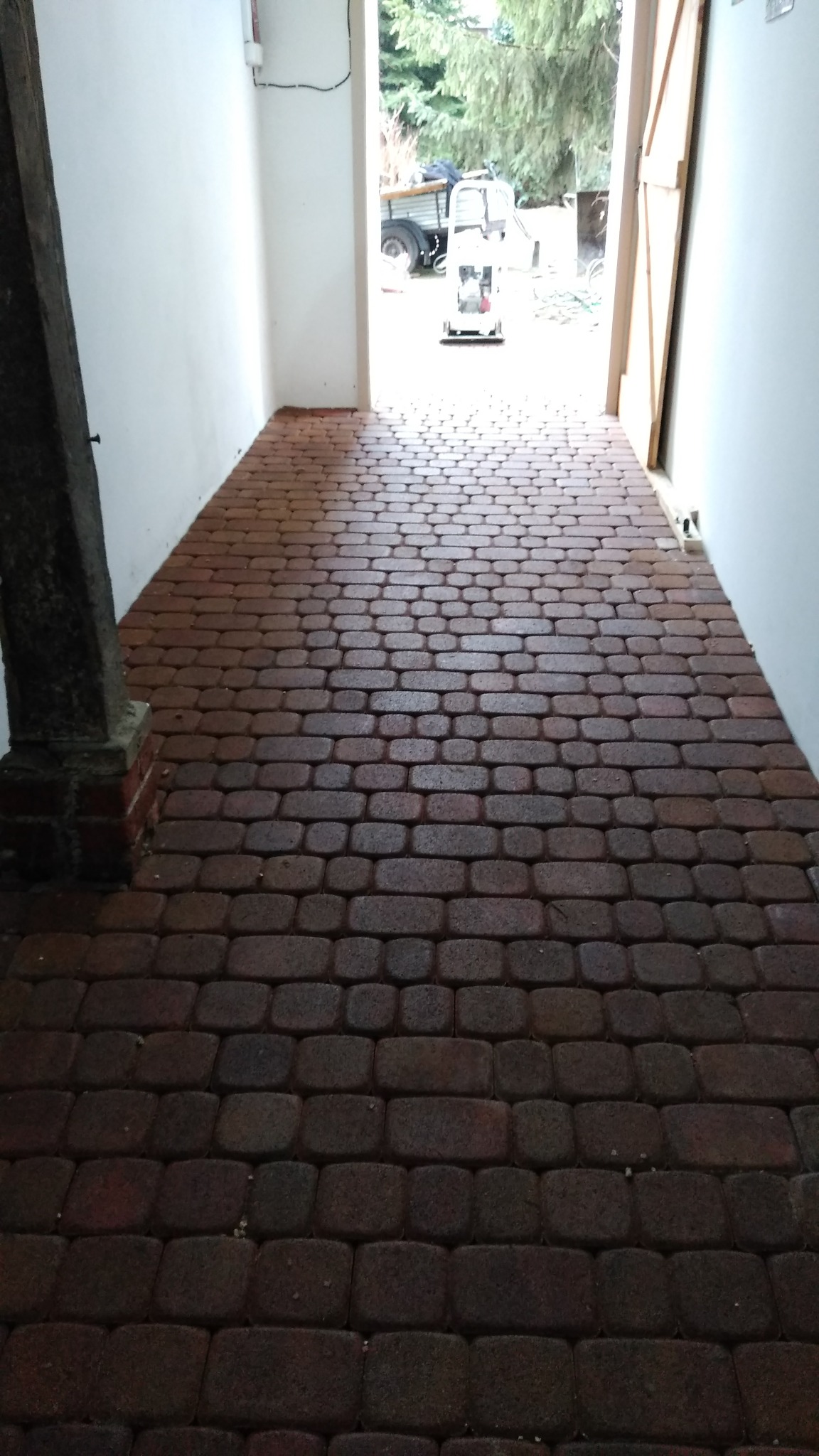 Nowo ułożona, mokra od deszczu, czerwono-brązowa kostka brukowa w przejściu między budynkami, z widoczną ubijarką w tle.