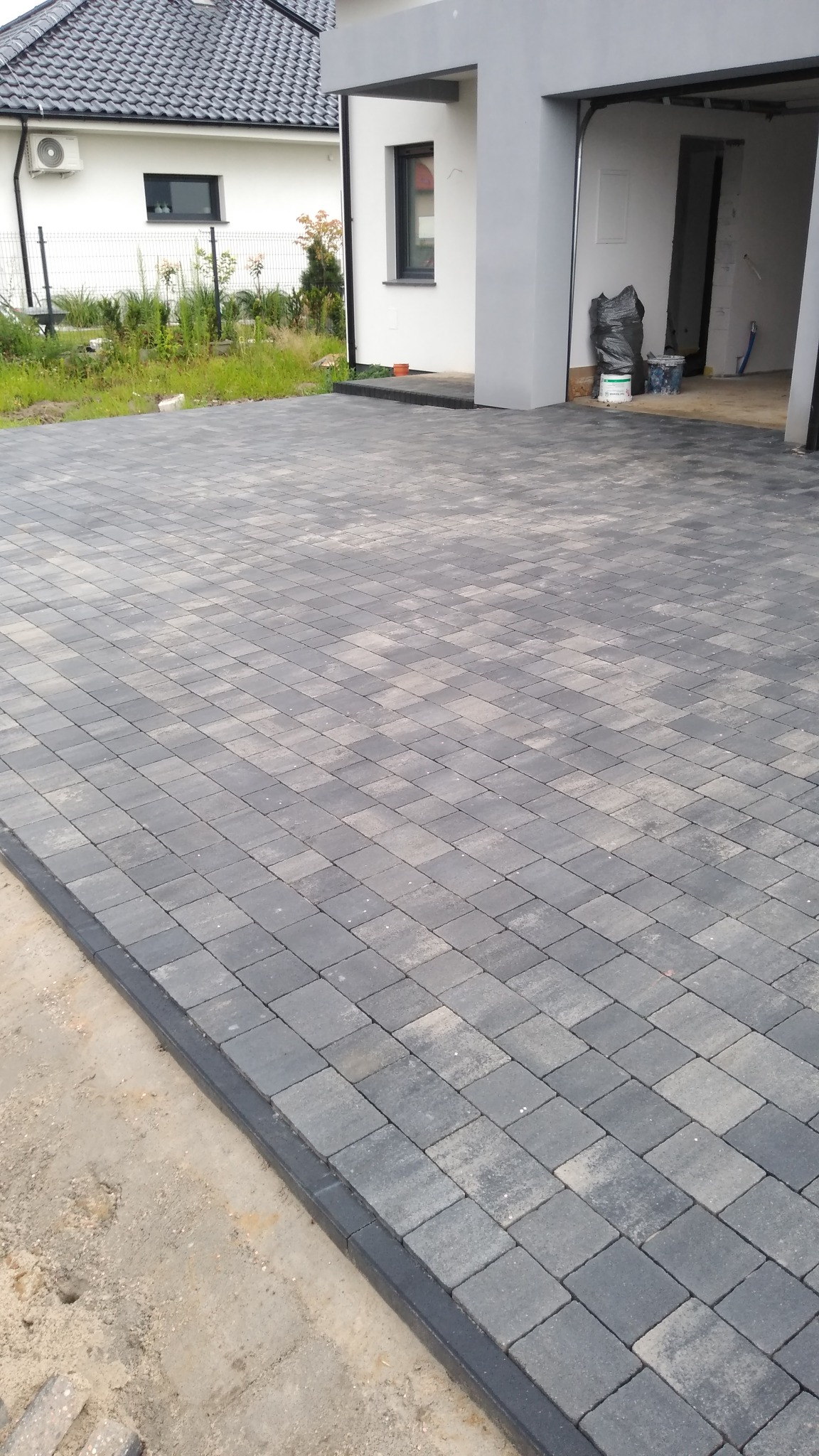 Ułożona, grafitowa kostka brukowa na podjeździe przed nowoczesnym domem z garażem, widoczne obrzeża i przygotowane podłoże po lewej stronie.