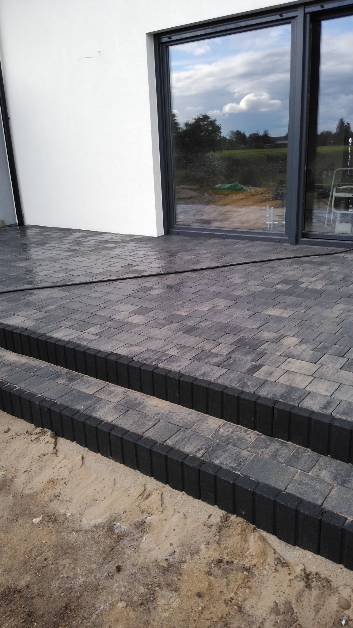 Nowo ułożony taras z kostki brukowej w odcieniach szarości z dwustopniowymi schodami, widoczne ślady piasku i odblaski wody po czyszczeniu, duże okno tarasowe odbija zachmurzone niebo i zieleń.