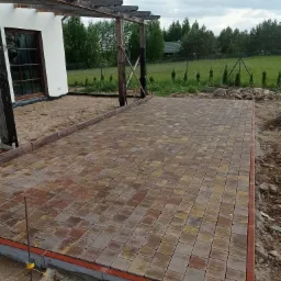 Ułożona kostka brukowa w odcieniach żółci i czerwieni, obrzeża z czerwonej kostki, widoczne narzędzia pomiarowe, w tle trawnik i drzewa.