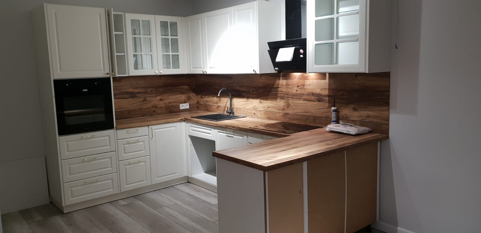Jasna kuchnia z białymi szafkami, drewnianym blatem i okładziną ścienną imitującą drewno, czarny zlew i okap, widoczne listwy przypodłogowe.