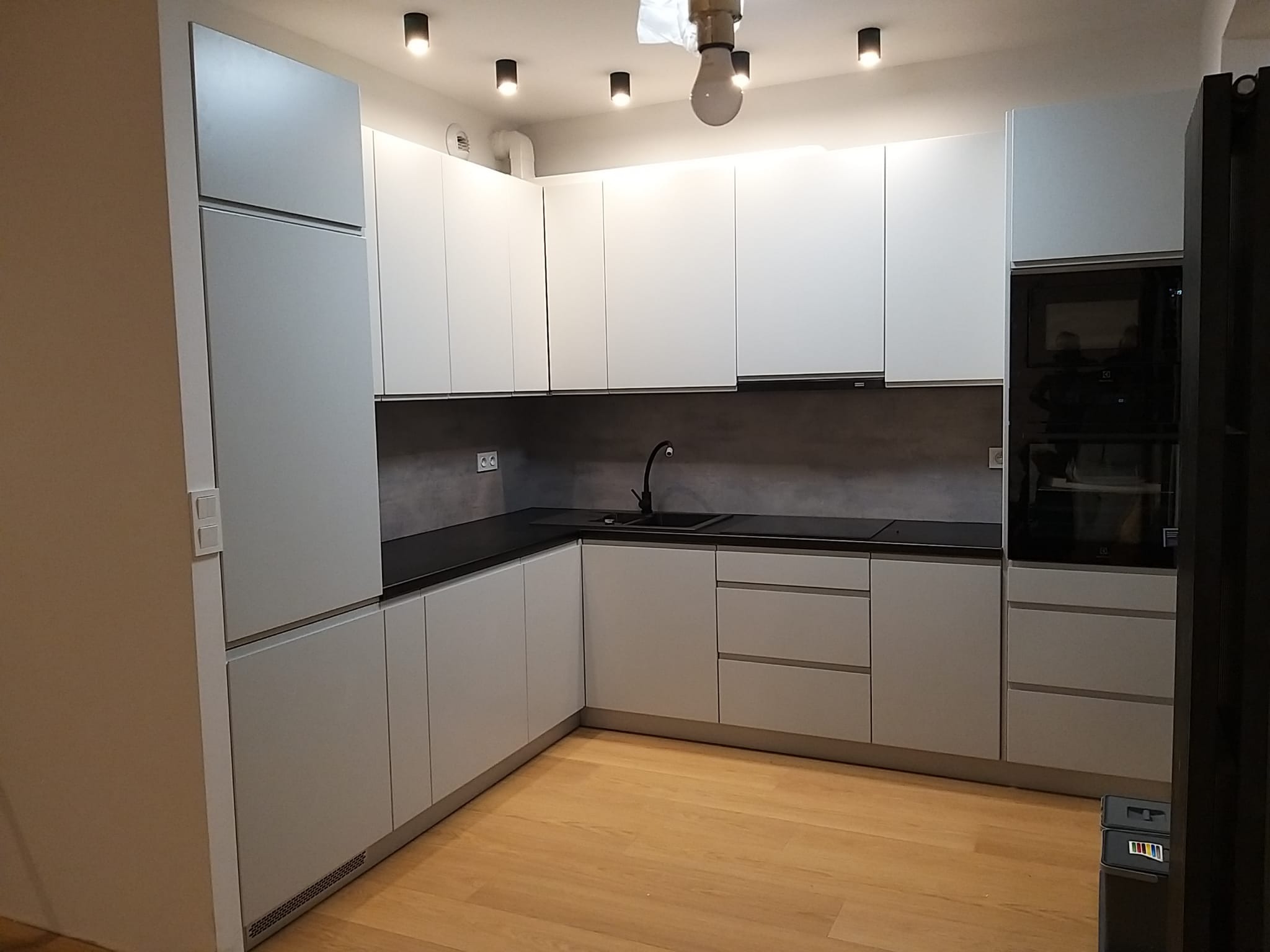 Nowoczesna, jasna kuchnia z białymi, matowymi frontami szafek i czarnym blatem, oświetlenie punktowe na suficie, parkiet na podłodze.